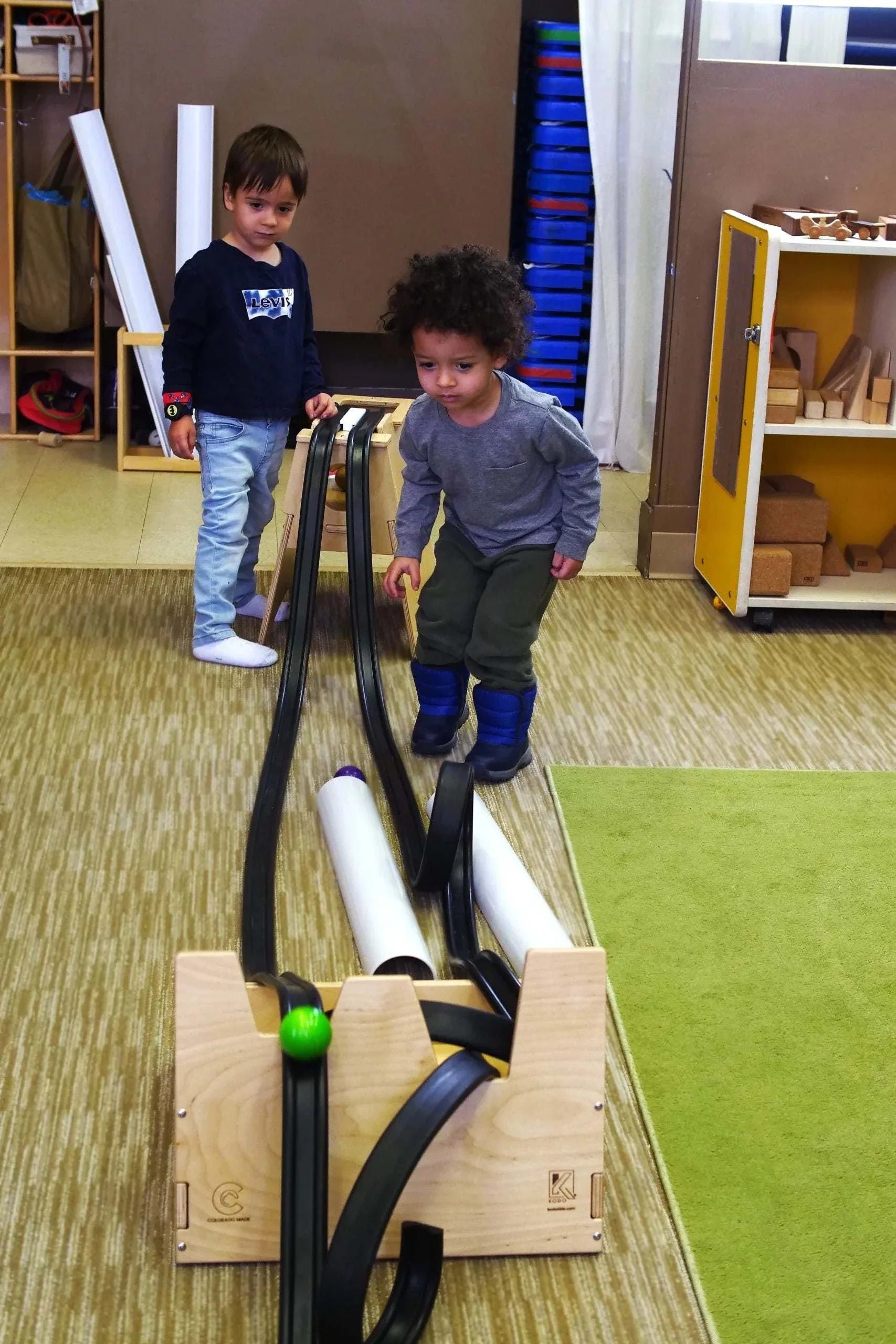 Two children playing with a wooden Rubber Ramp Tower set featuring flexible black tracks and colorful balls on a classroom floor.