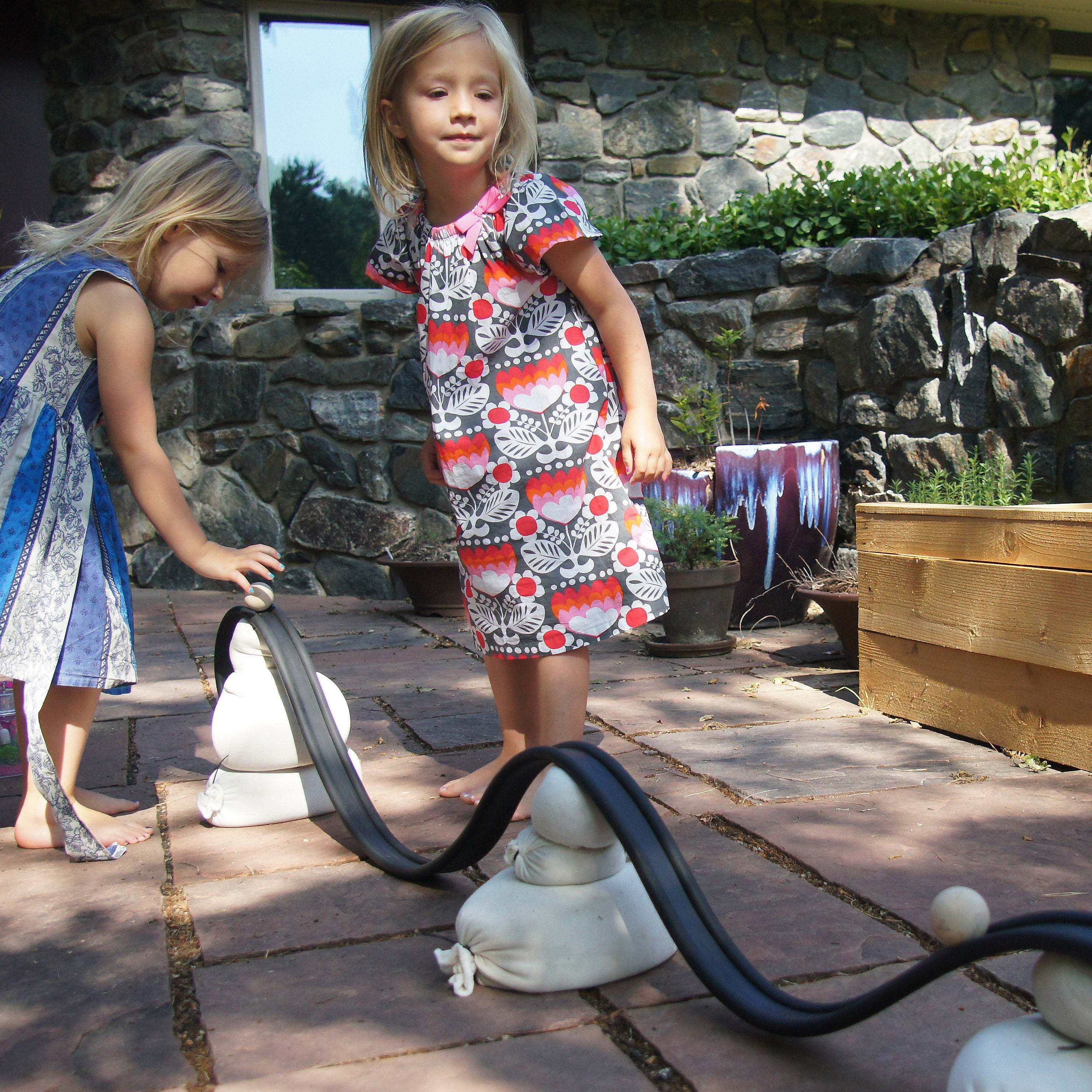 Two girls playing with a flexible black rubber ramp supported by white sandbags on a stone patio.