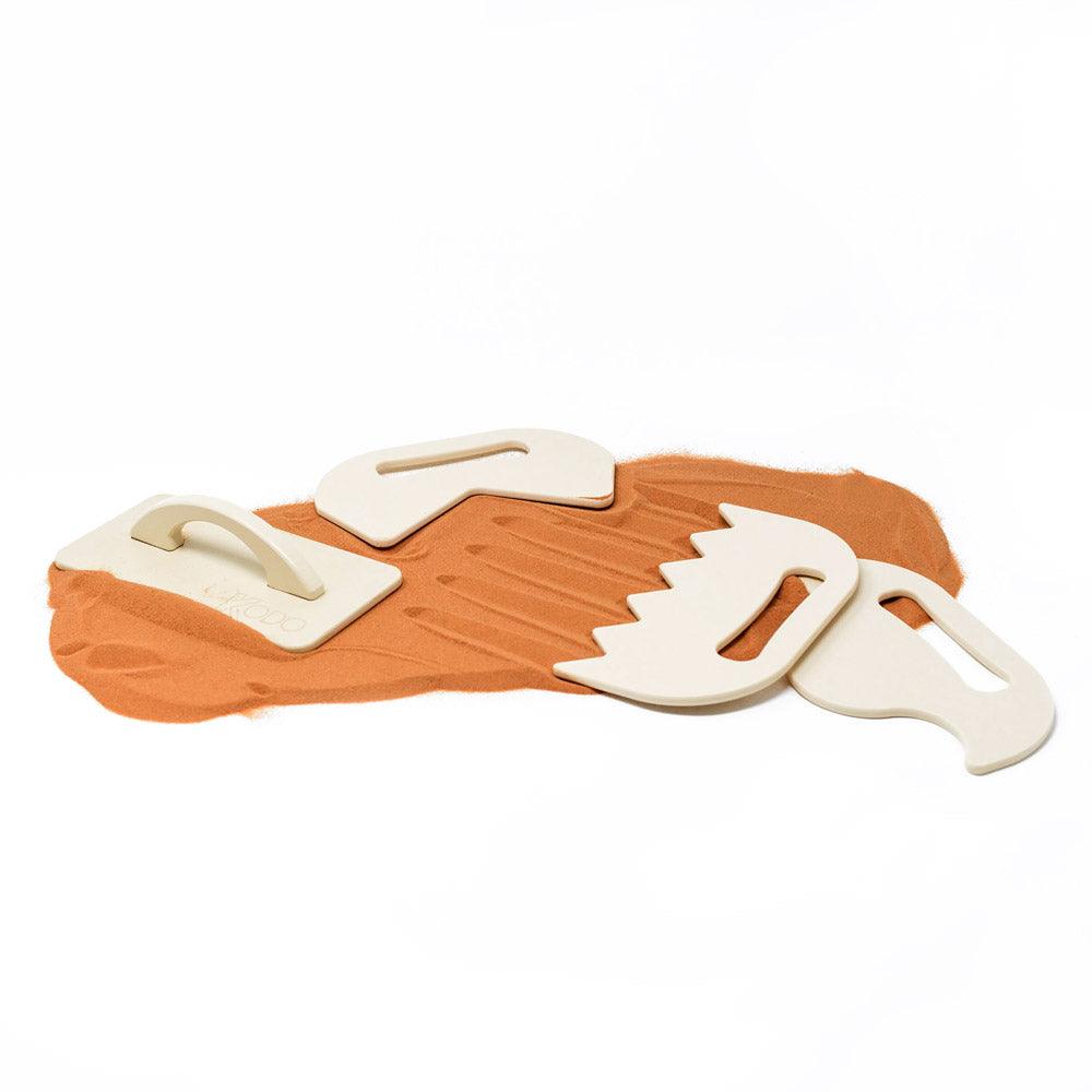 Four beige plastic sand shaping tools with handles resting on a pile of orange sand against a white background.