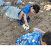 Child using a white, jagged-edged plastic Sand Shaper tool to rake patterns in a sandy outdoor play area.