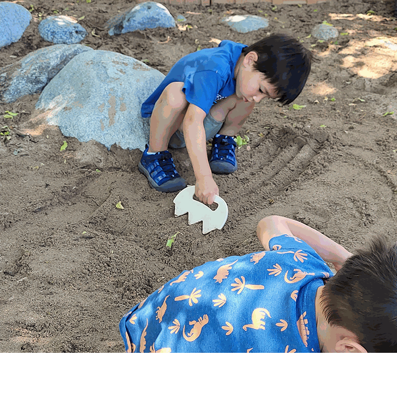 Child using a white, jagged-edged plastic Sand Shaper tool to rake patterns in a sandy outdoor play area.