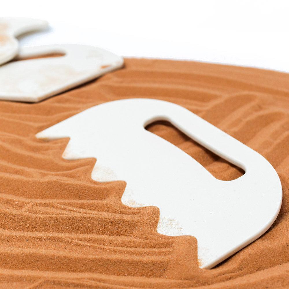 White plastic sand shaping tool with a serrated edge resting on textured brown sand.