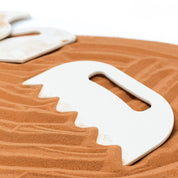 White plastic sand shaping tool with a serrated edge resting on textured brown sand.