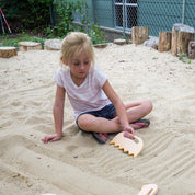 Young girl using a light-colored wooden Sand Shaper tool to create textured patterns in a backyard sandbox.