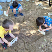 Children using white, ergonomic plastic Sand Shapers to create patterns and textures in a sandy outdoor play area.