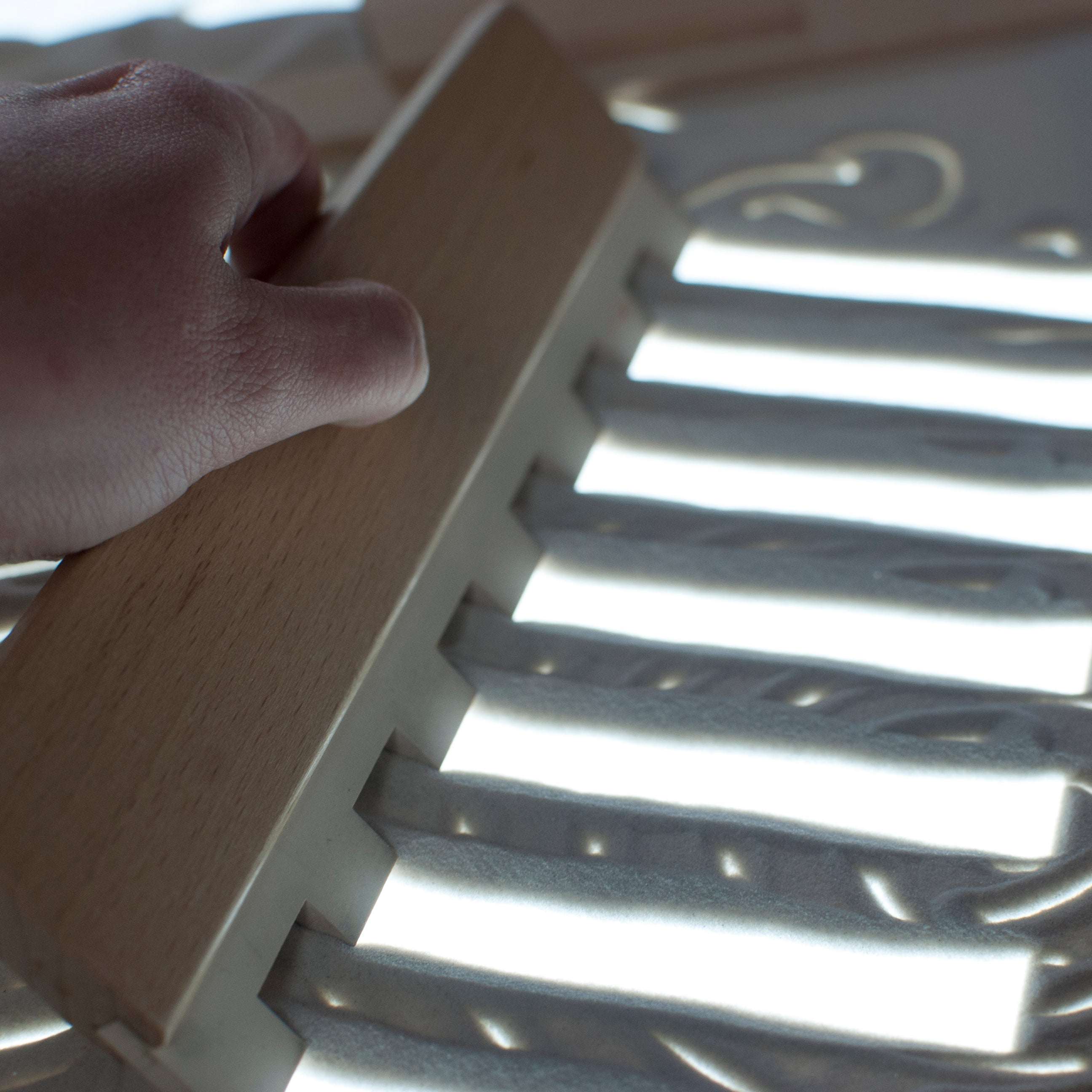 Hand using a wooden rake tool on a bright, rectangular LED light panel for sensory sand play.