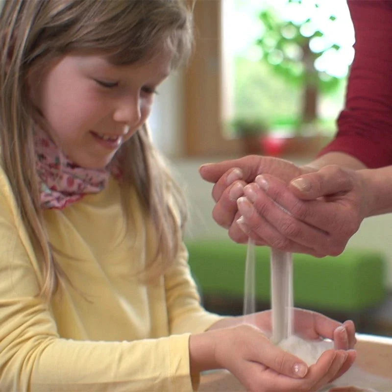 A young girl watches as fine, silvery sand pours from an adult's hands into her own for sensory sand tray play.