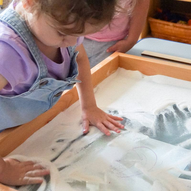 Child using fine, silvery sand in a wooden tray to practice drawing patterns and shapes.