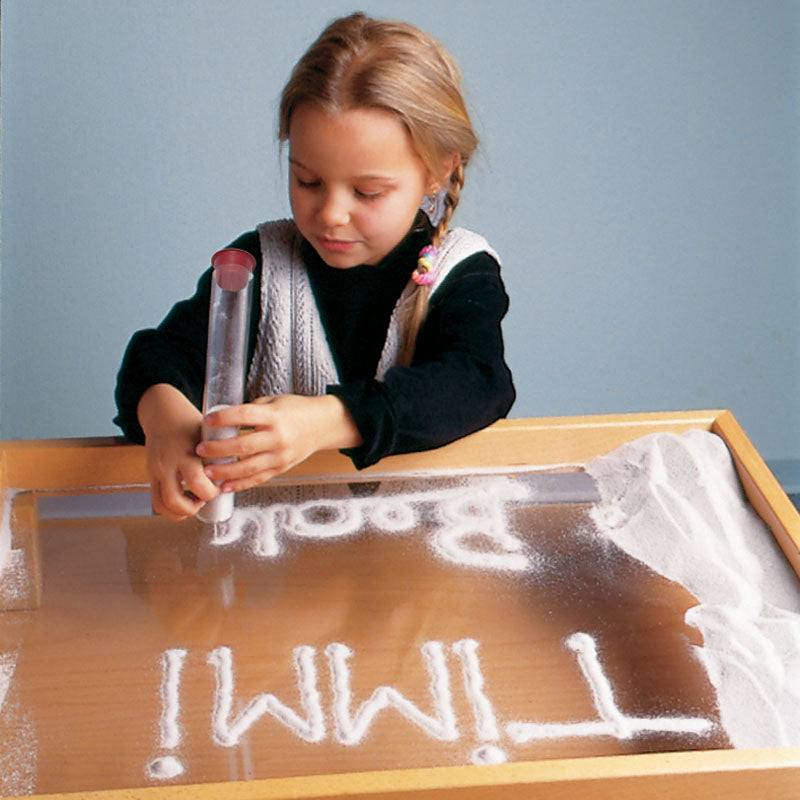 A child using a clear, funnel-shaped Sand Writer pen to draw letters in white sand on a wooden sensory tray.
