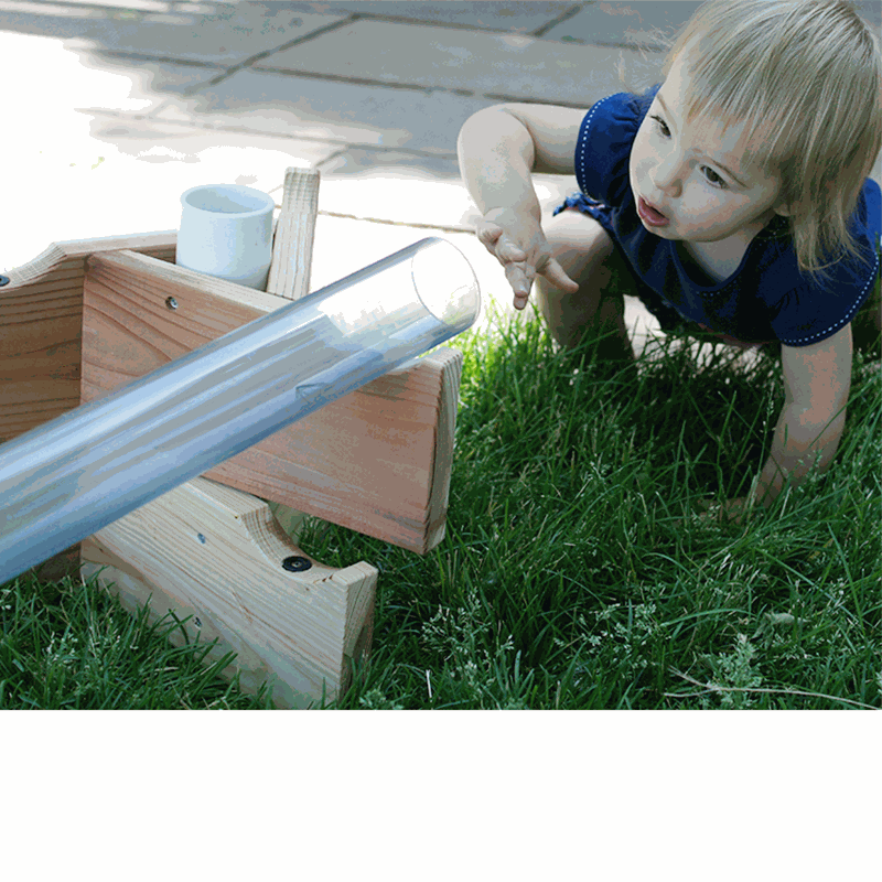Child playing with a clear, durable plastic pipe mounted on a wooden stand for outdoor STEM sand and water exploration.