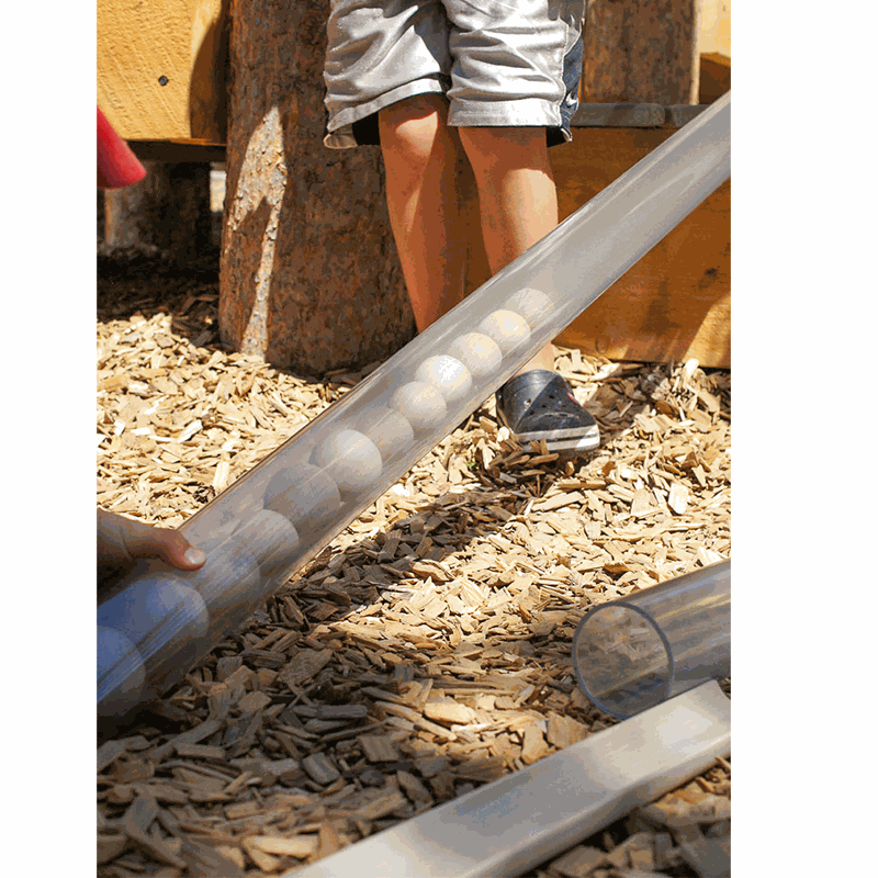 Transparent plastic pipe filled with wooden balls for STEM play, resting on a wood chip outdoor surface.