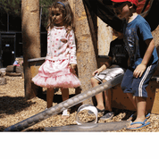 Children playing with clear, cylindrical See Inside Pipes filled with wooden balls in an outdoor STEM learning environment.
