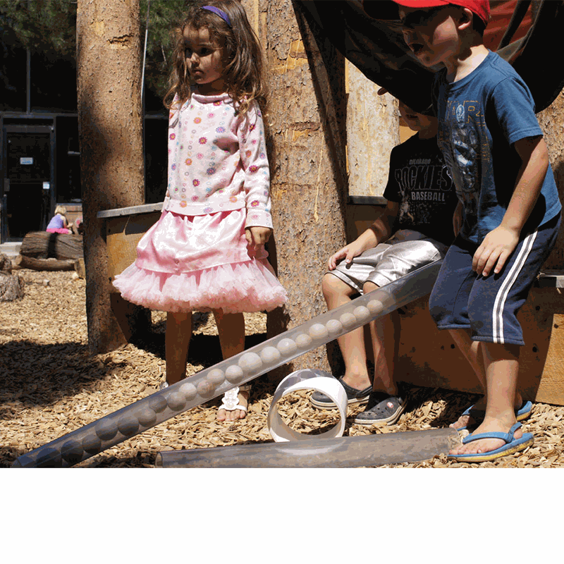 Children playing with clear, cylindrical See Inside Pipes filled with wooden balls in an outdoor STEM learning environment.
