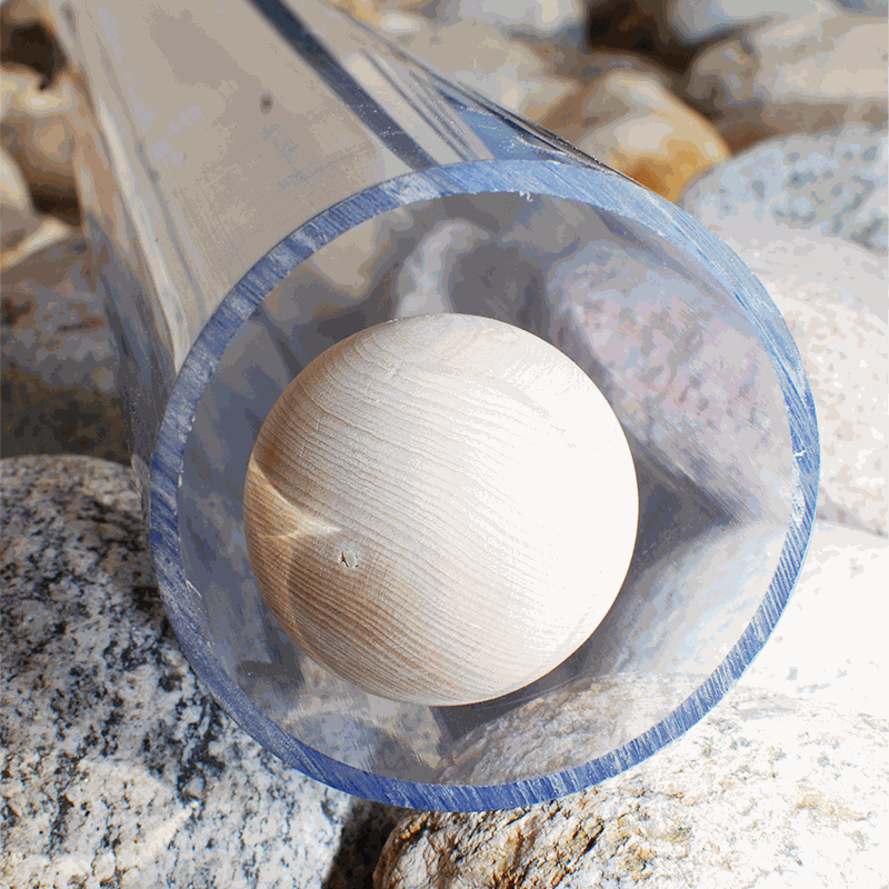 A smooth wooden ball rolls through a clear, durable plastic pipe resting on a bed of grey river stones.