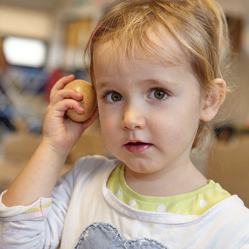 A young child holding a realistic, speckled sensory sound egg to her ear to listen to the auditory feedback.