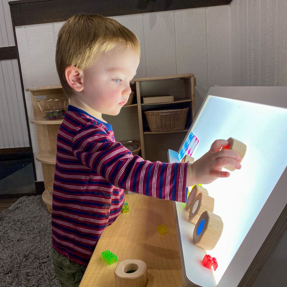 Child playing with wooden and transparent blocks on an upright, glowing white shelf-top light panel in a classroom.