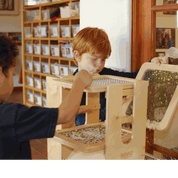 Children using a light-colored wooden Sift & Sort tower with three removable mesh screens to filter sensory materials.