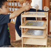 Children using a three-tiered wooden Sift & Sort tower with removable mesh screens to classify sensory materials by size.