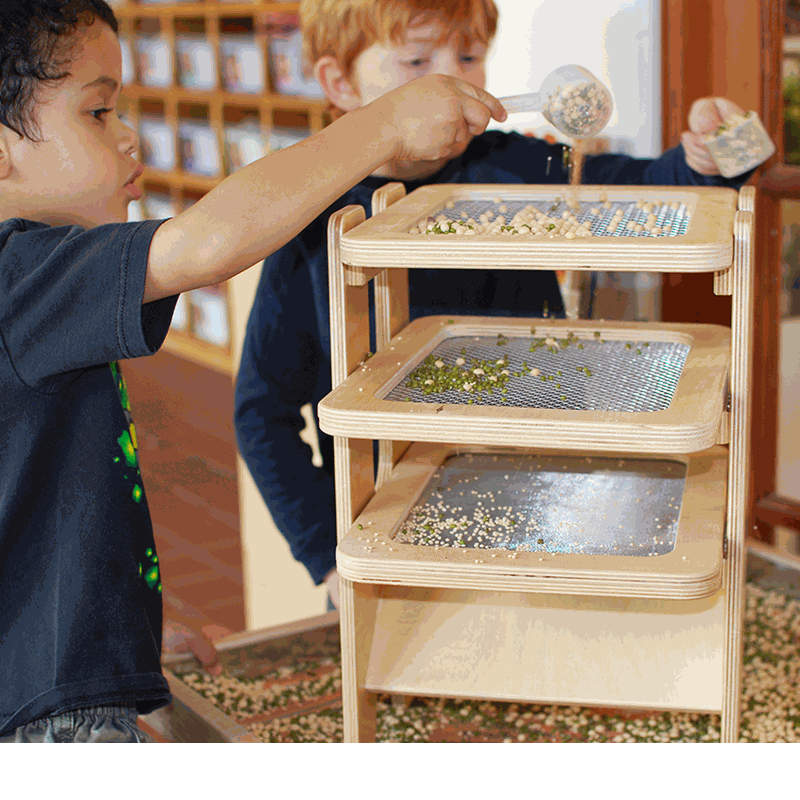 Children using a three-tiered wooden Sift & Sort tower with removable mesh screens to classify sensory materials by size.