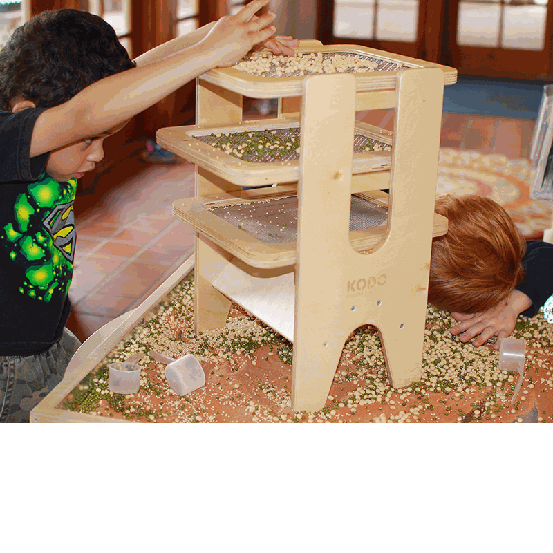 Children using a wooden Sift & Sort tower with three removable mesh screens to classify sensory materials by size.