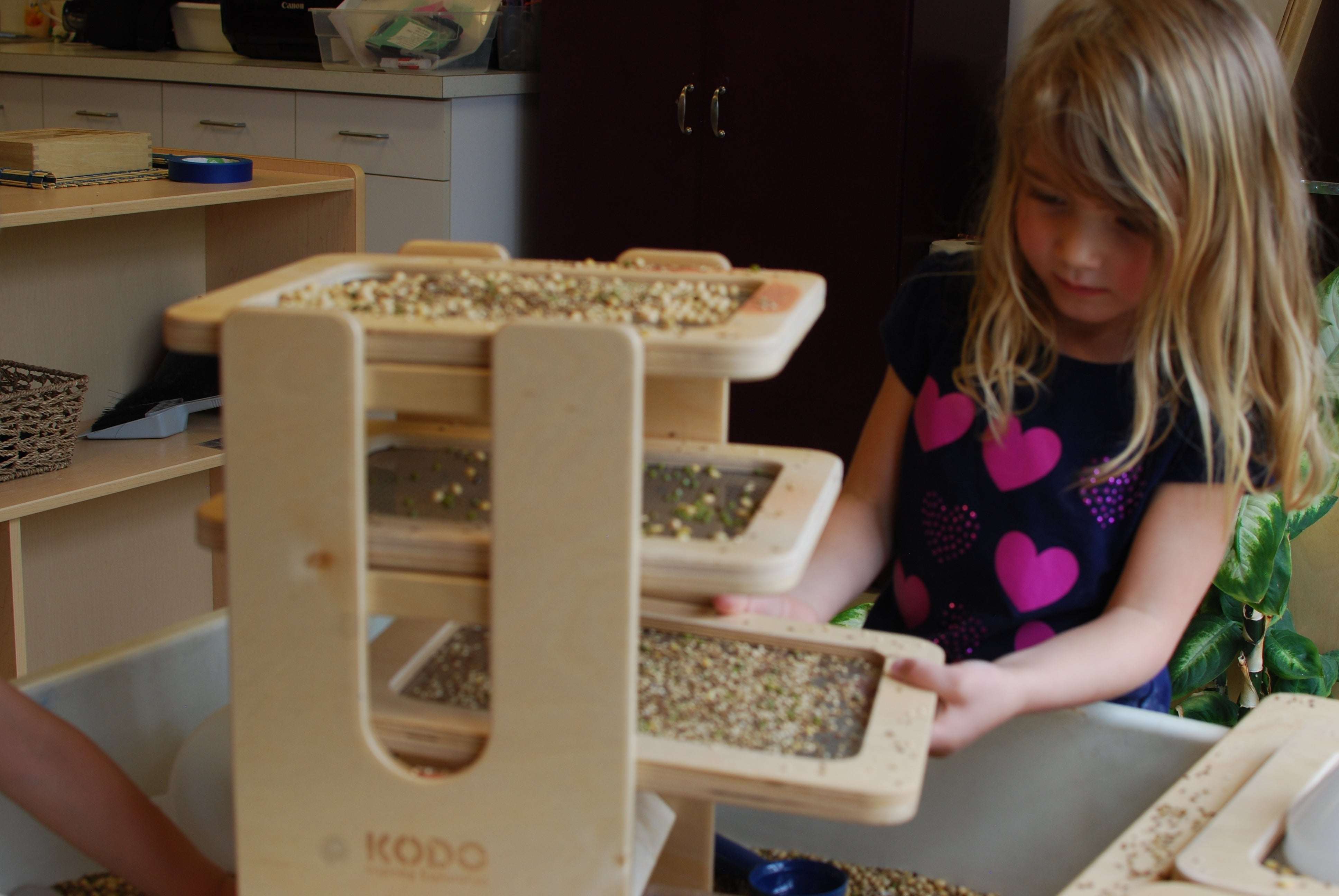 Child using a wooden Sift & Sort tower with three removable mesh screens to classify sensory materials by size.