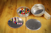 Two stackable metal bowls and two flat plates on a wooden floor, filled with colorful pom-poms and wooden pegs.