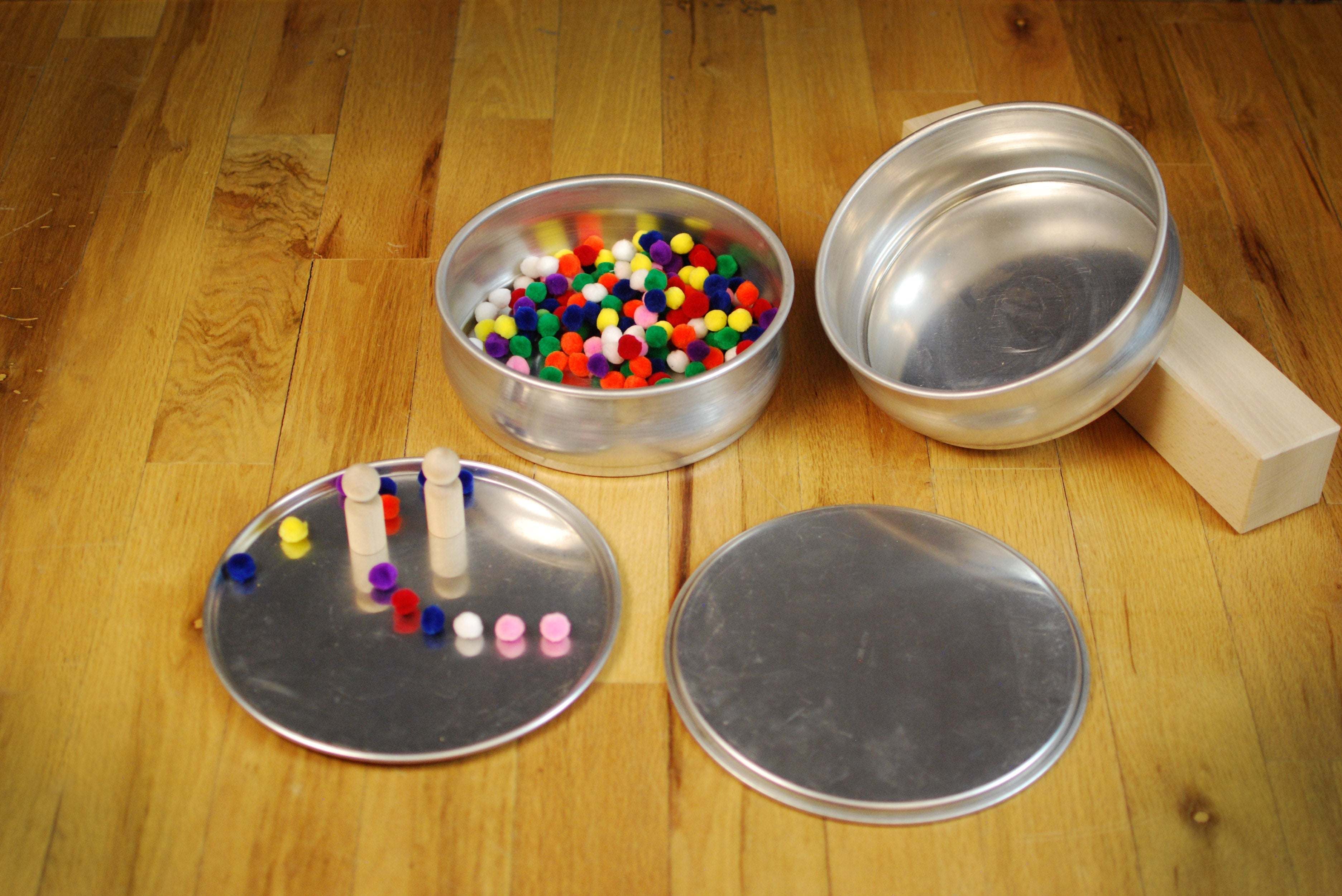 Two stackable metal bowls and two flat plates on a wooden floor, filled with colorful pom-poms and wooden pegs.