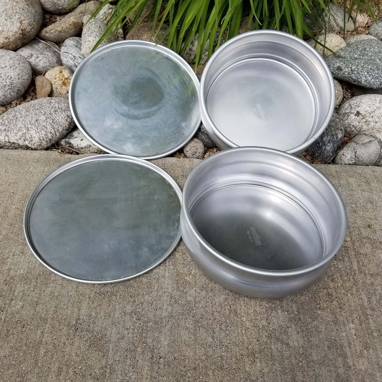 Two silver metal bowls and two matching flat plates arranged on a concrete surface outdoors.