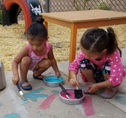 Two children using small, round, silver metal pans filled with colorful paint and brushes for outdoor art activities.