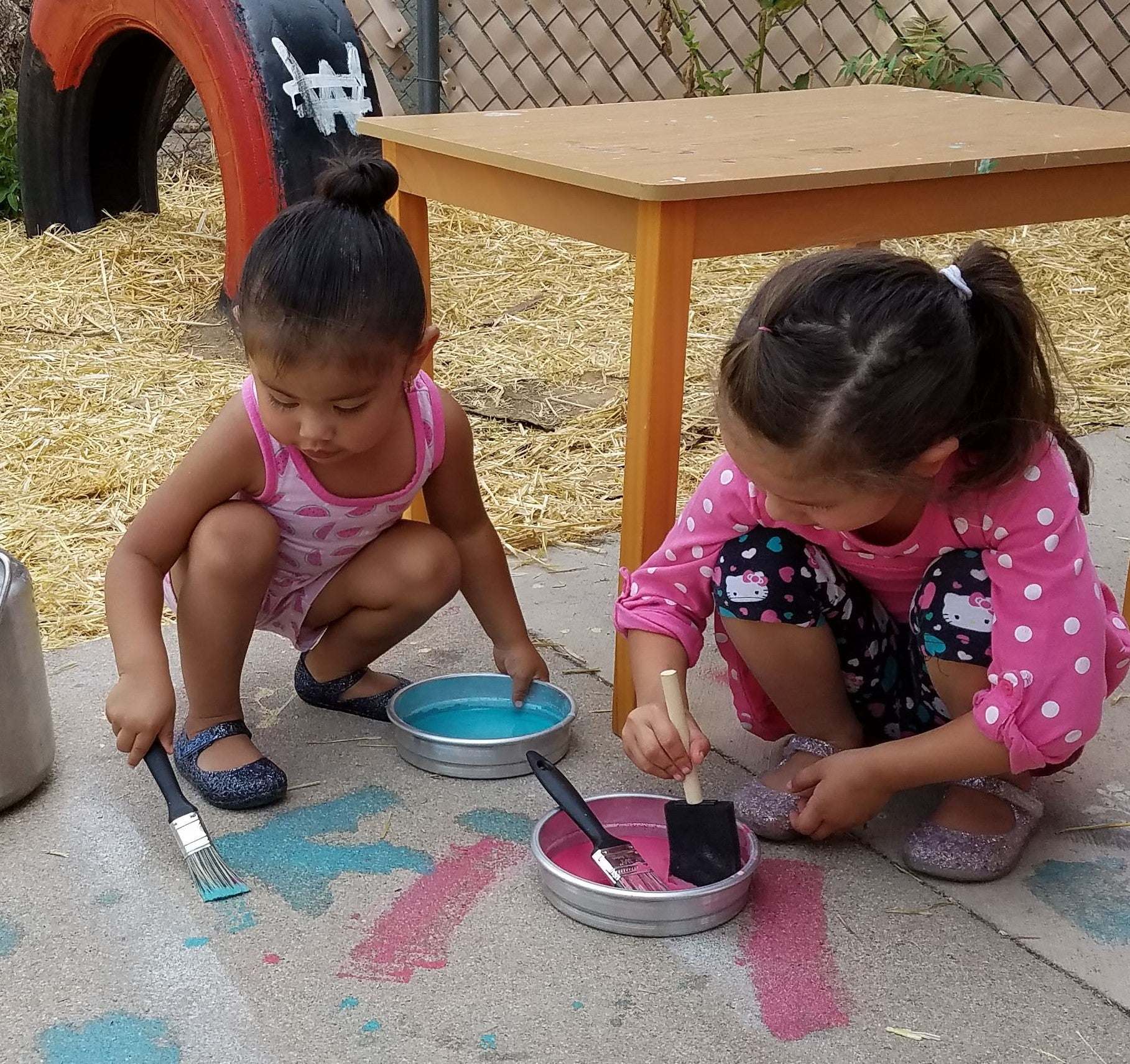 Two children using small, round, silver metal pans filled with colorful paint and brushes for outdoor art activities.