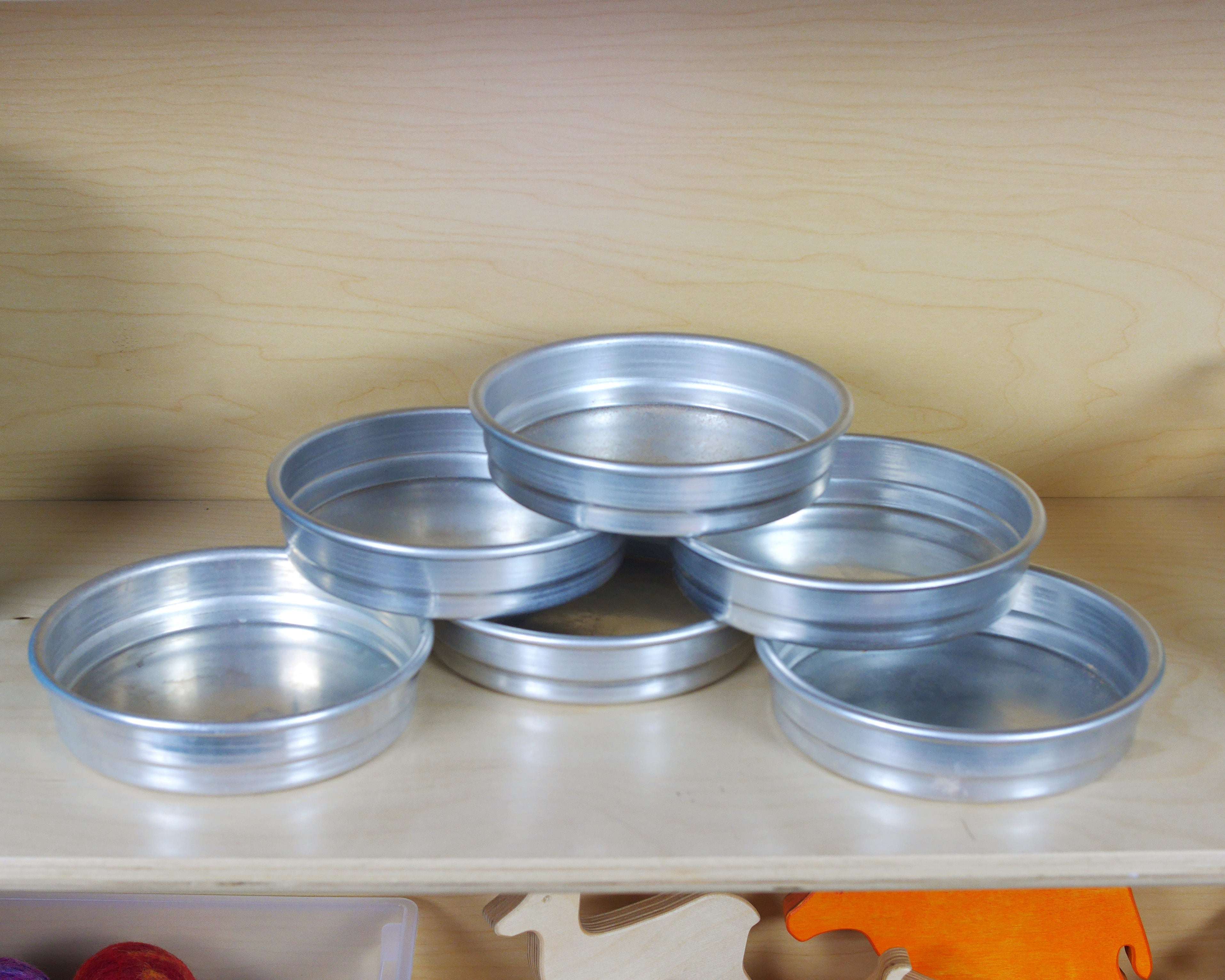 Set of five stackable, round aluminum pans for children's play, arranged on a wooden shelf.