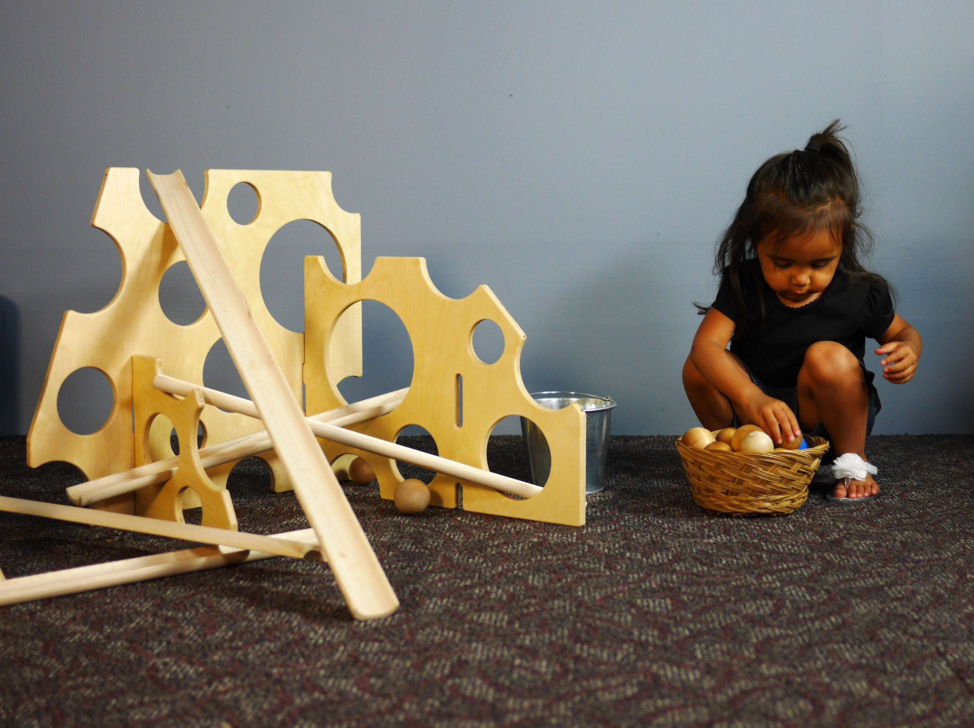 Child playing with light wood Swiss cheese-style ramp stands and wooden balls on a carpeted floor.