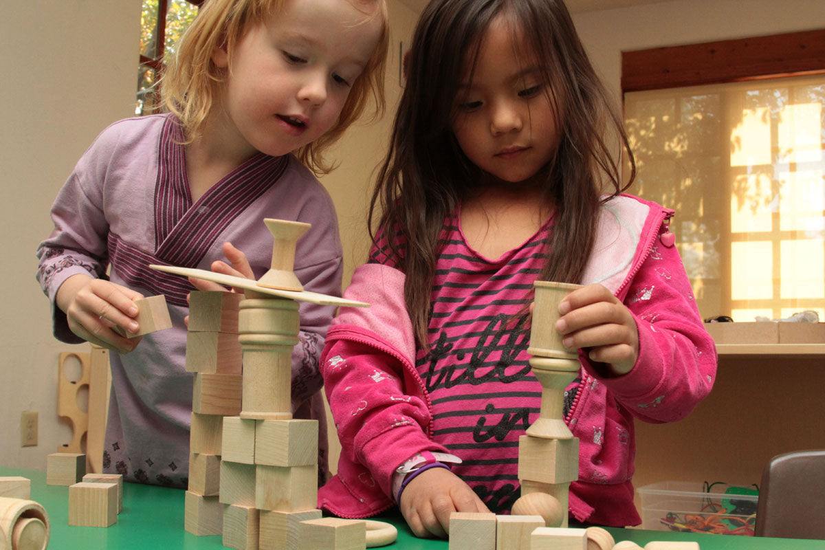 Two children building structures with smooth, natural wooden cubes and rings on a green tabletop.
