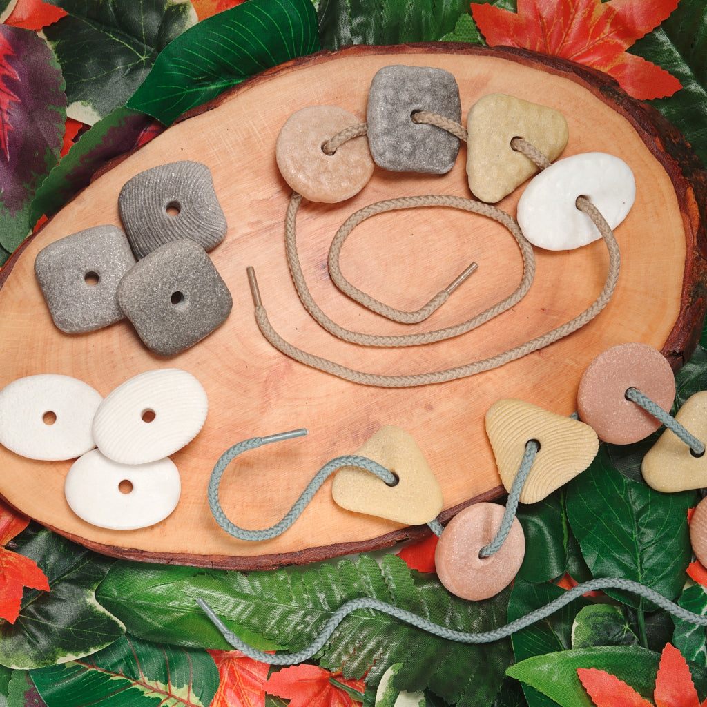 Set of 16 tactile threading pebbles in various shapes and colors with threading cords on a wooden slice background.