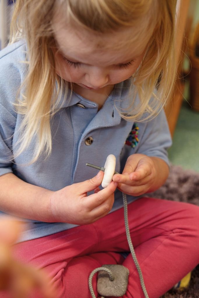 A young child threading a smooth, white tactile pebble onto a grey cord to develop fine motor skills.