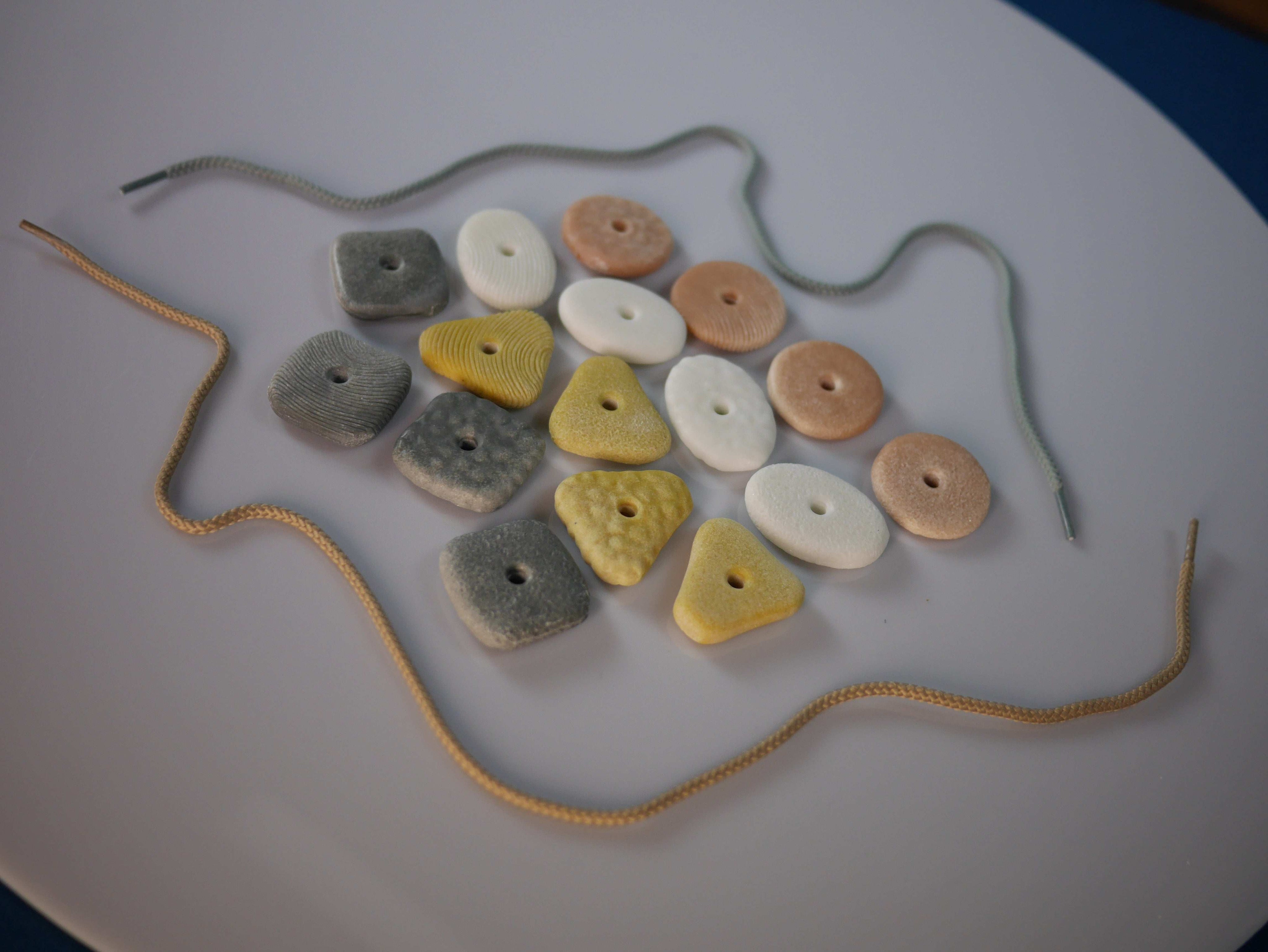 Set of 16 tactile threading pebbles in assorted shapes, colors, and textures with two threading laces on a white surface.