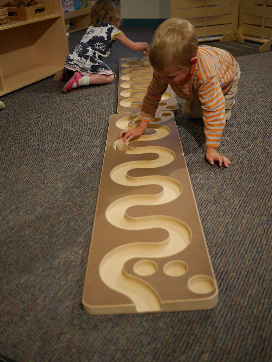Toddlers playing with a wooden ball run featuring a winding, carved track on a carpeted floor.