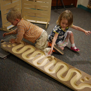 Two toddlers playing with a wooden ball run featuring a winding, serpentine track on a carpeted floor.