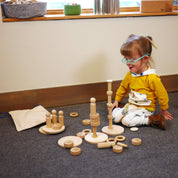 Toddler playing with natural wooden stacking blocks, rings, and figures on a gray carpet in a playroom.