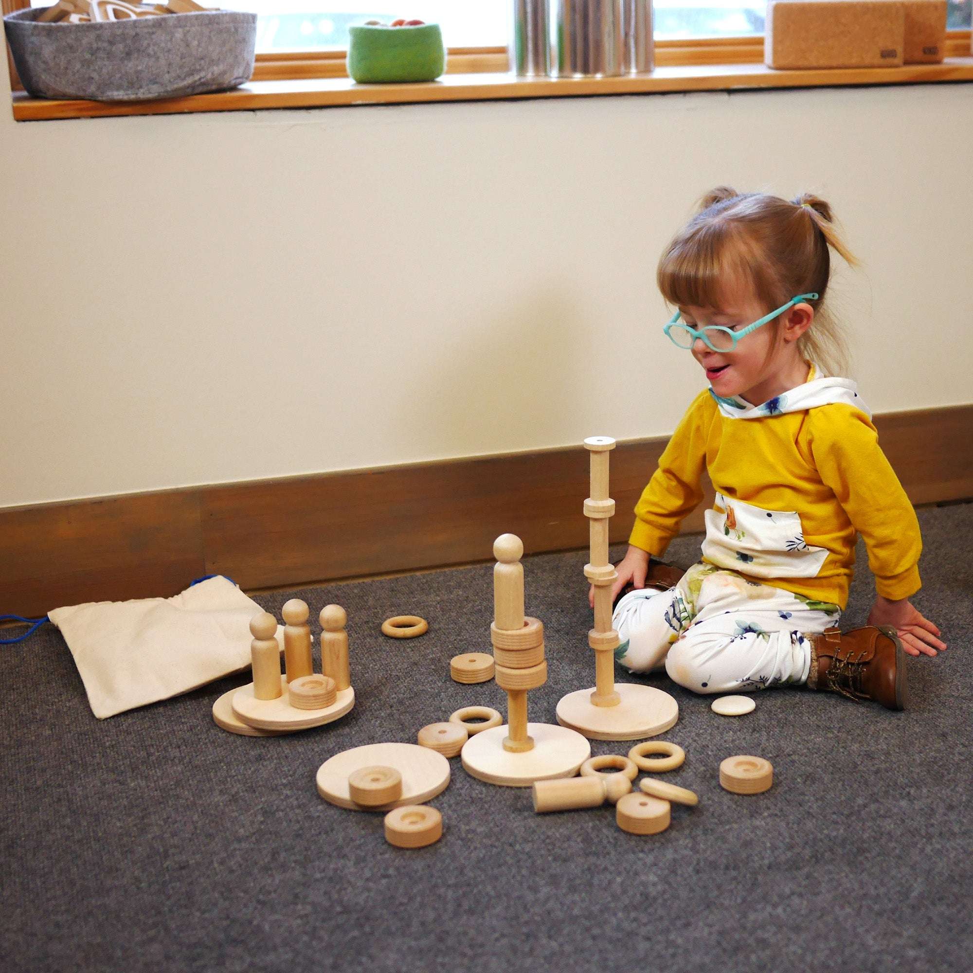 Toddler playing with natural wooden stacking blocks, rings, and figures on a gray carpet in a playroom.