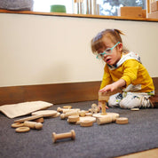 Toddler playing with natural wooden building blocks, wheels, and spools on a grey carpet.