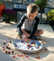 Child playing with translucent white sensory beads and nature items in a round metal tray on a textured mat.