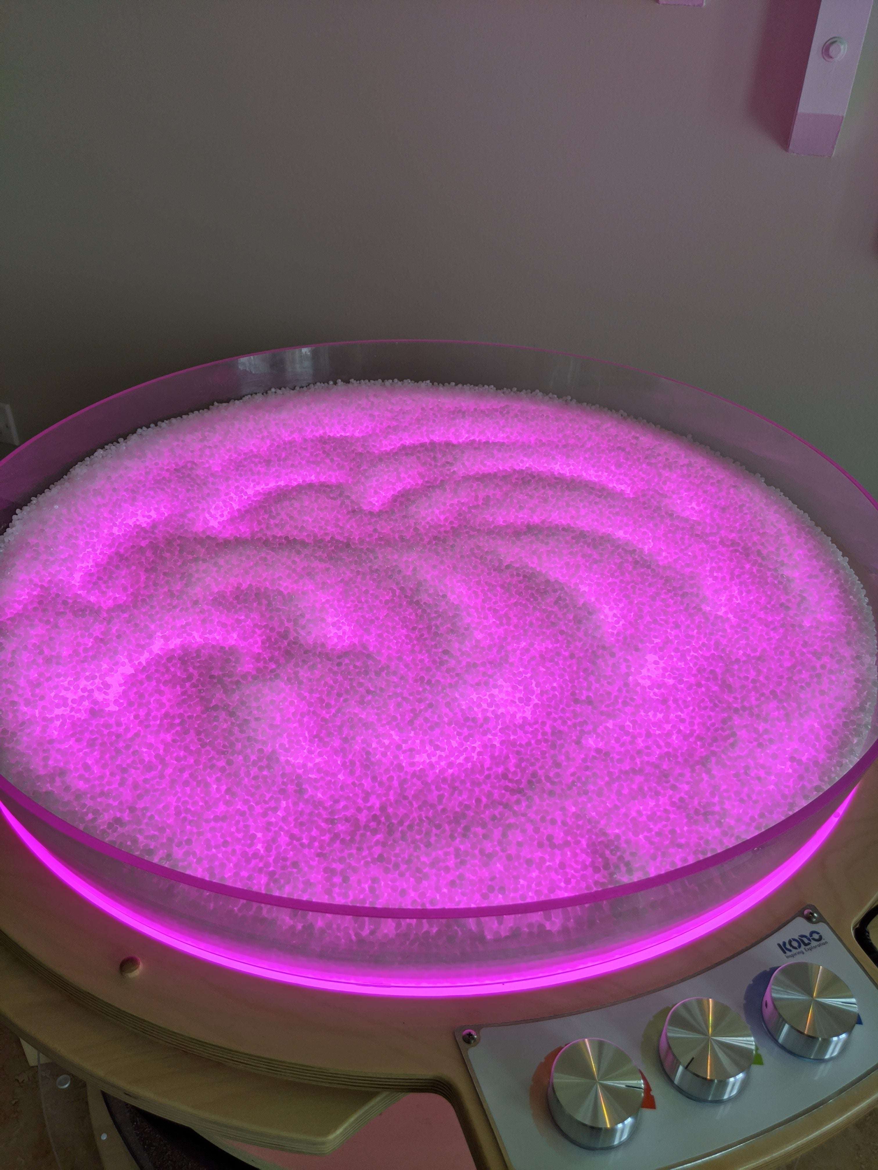Milky white translucent sensory beads glowing with vibrant pink light in a circular Kodo Messy Materials tray.