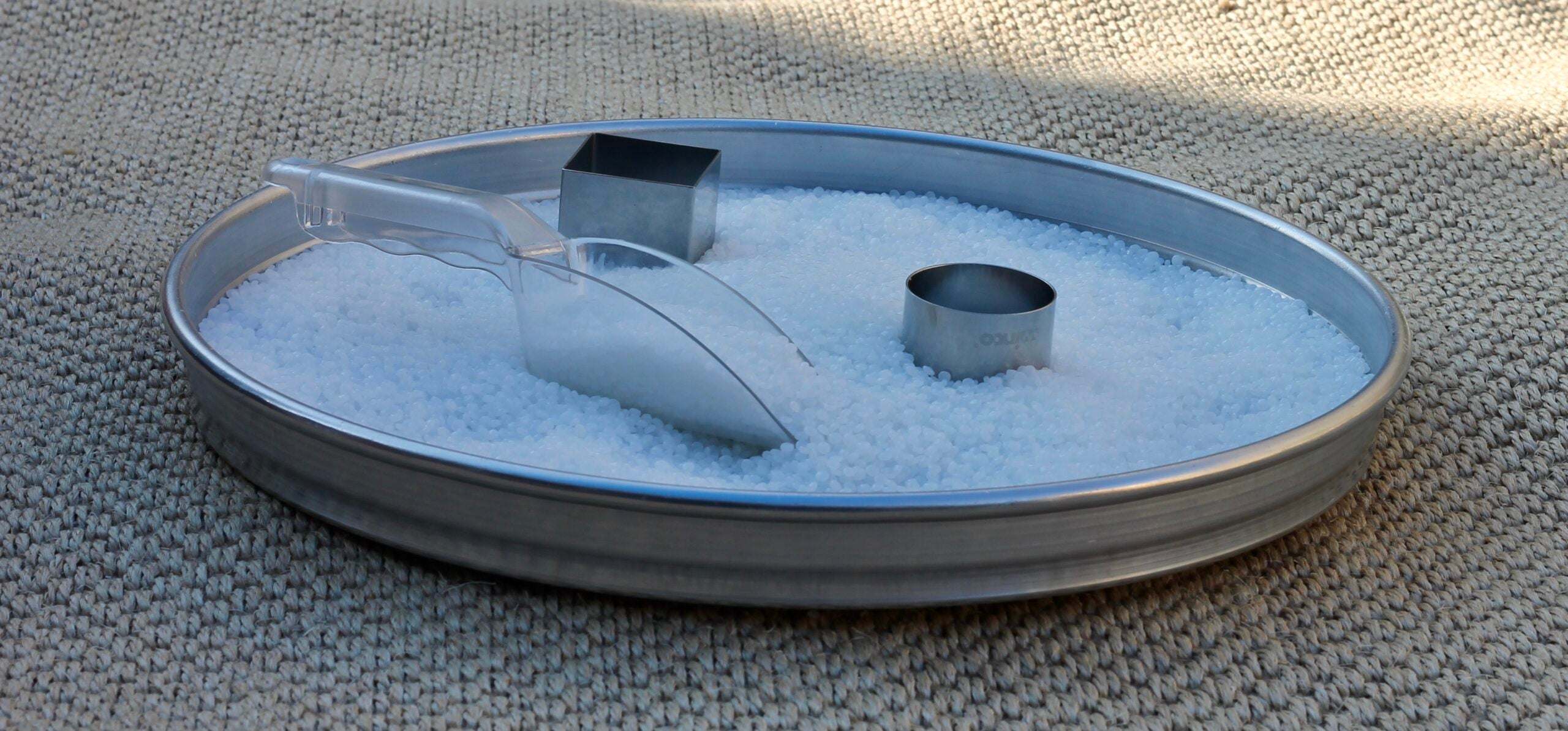 Milky white translucent sensory beads in a metal tray with a clear plastic scoop and two metal shape molds.