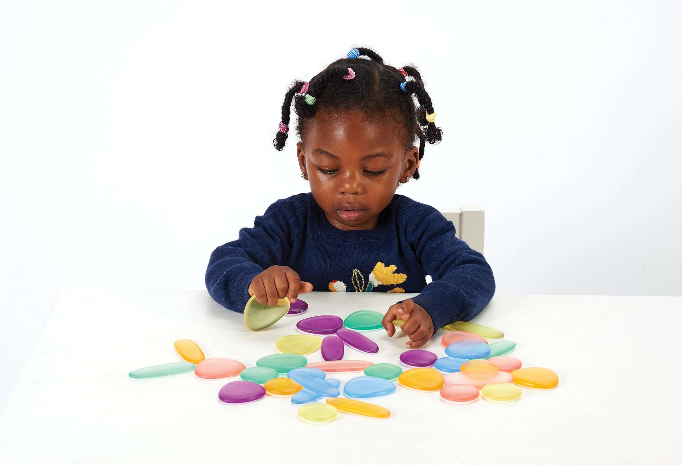 Young child playing with colorful, transparent, smooth-edged pebbles of various sizes on a white table.