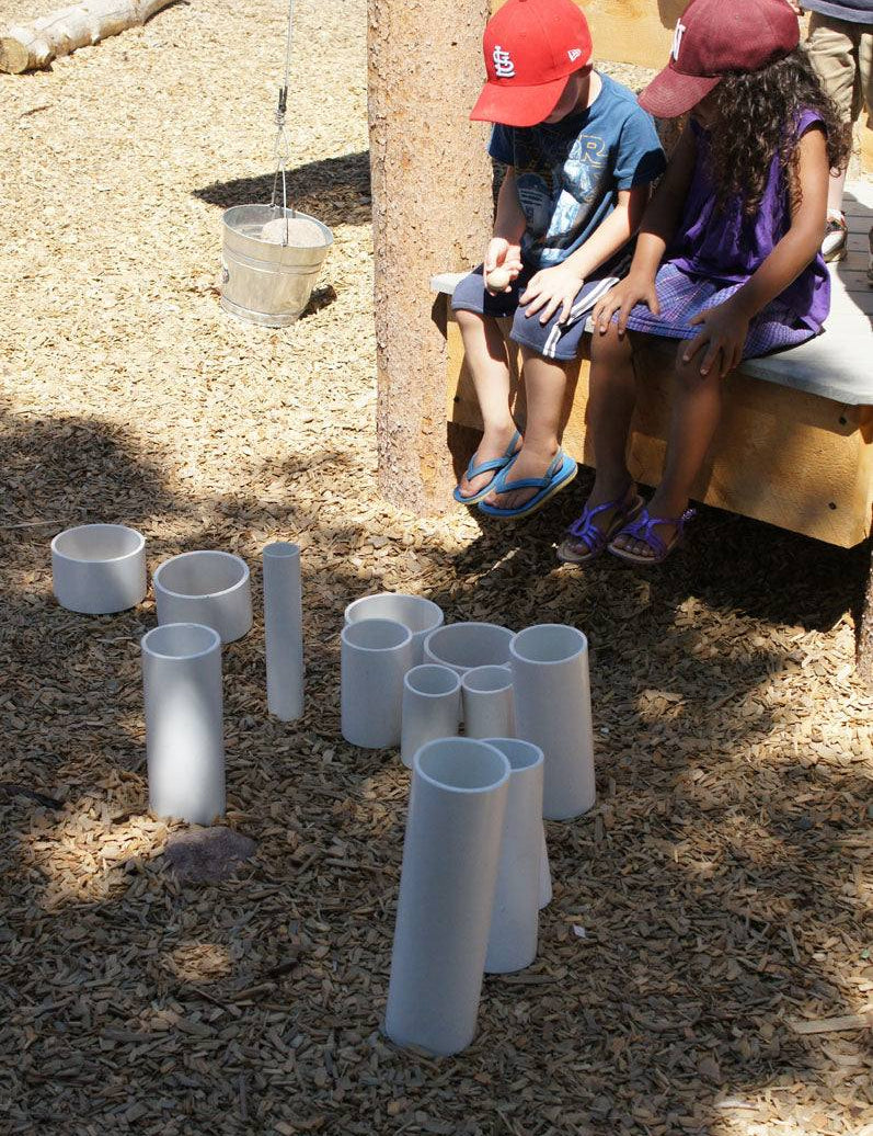 Children playing with a set of rigid white PVC tubes of varying lengths arranged upright on a wood chip playground surface.