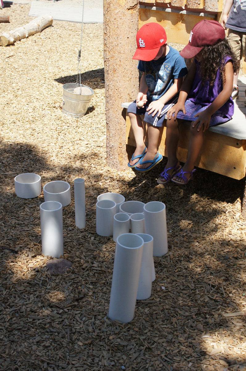 Children playing with a set of rigid white PVC tubes of varying lengths arranged upright on a wood chip playground surface.