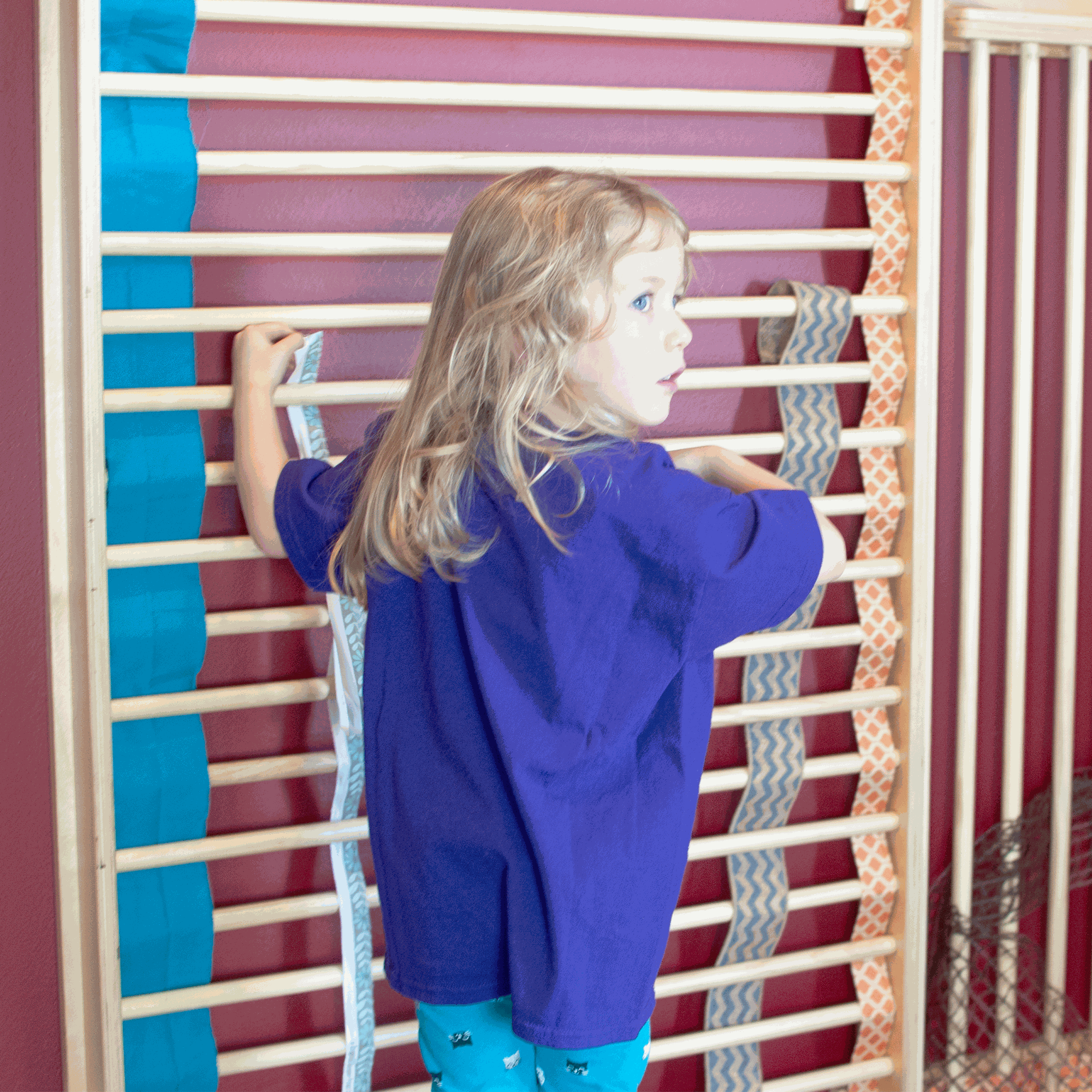 Young girl in a purple shirt climbing a tall, light wood wall-mounted ladder with colorful fabric ribbons attached.