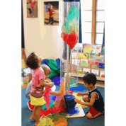 Children playing with a tall, clear pivoting wind tunnel tube and colorful play silks in a classroom setting.