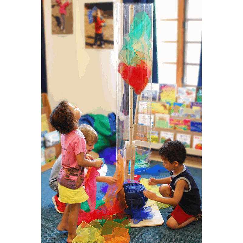Children playing with a tall, clear pivoting wind tunnel tube and colorful play silks in a classroom setting.